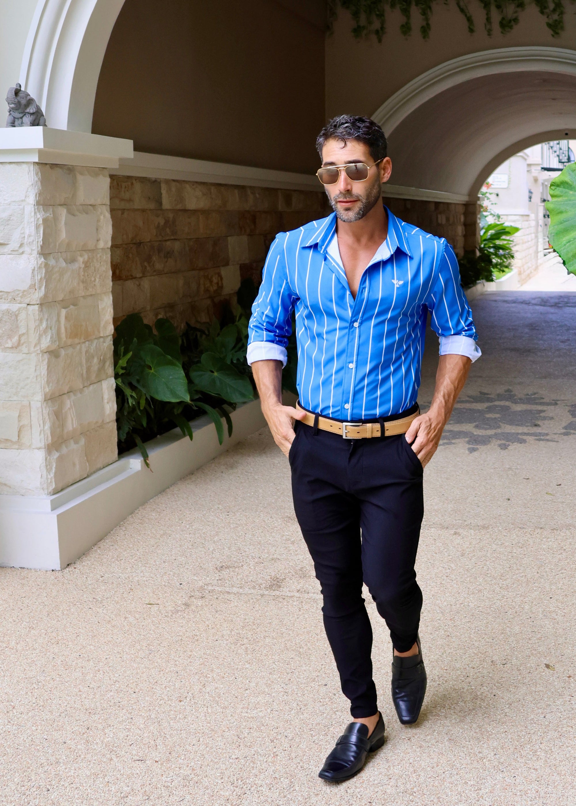 Man in a blue striped shirt and black pants standing in an outdoor setting with stone walls and plants.
