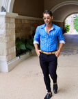 Man in a blue striped shirt and black pants standing in an outdoor setting with stone walls and plants.