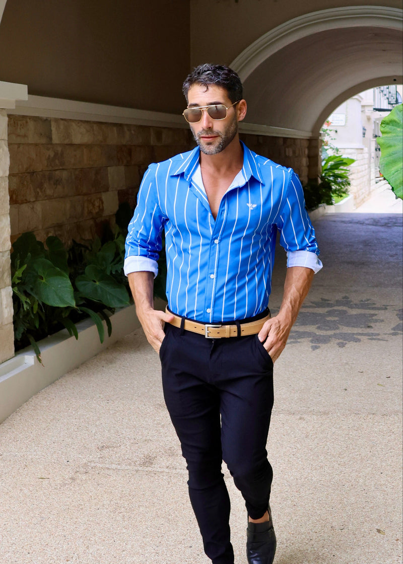Man in a blue striped shirt and black pants standing in an outdoor setting with stone walls and plants.